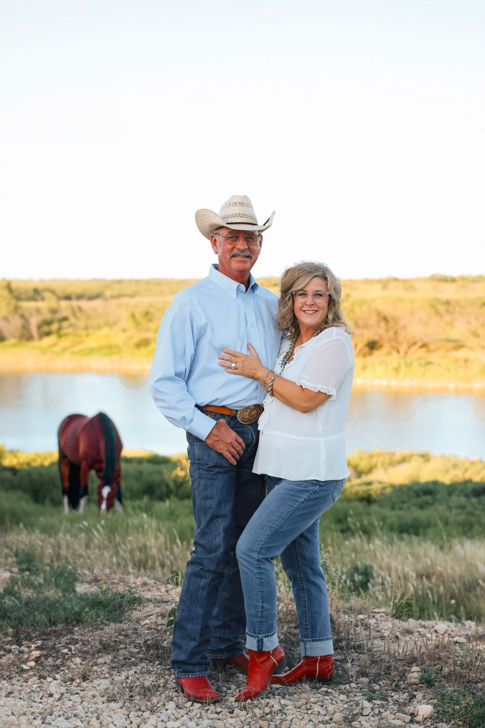 Couple in western attire posing together by a lake with red boots and casual denim.