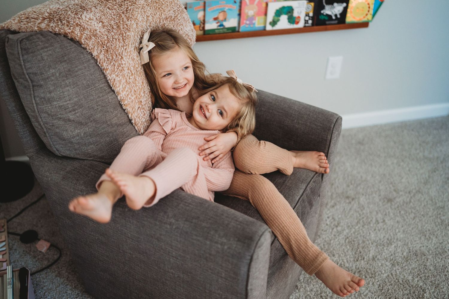 Family having fun and cuddling together on a cozy grey sofa with children's books displayed on wall shelves.