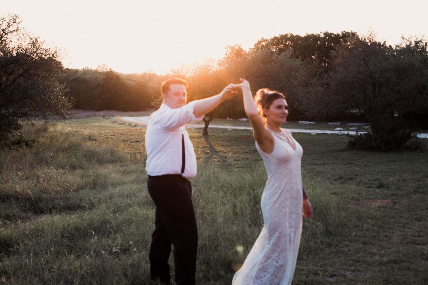 Dancing couple in wedding attire silhouetted against golden sunset in field.