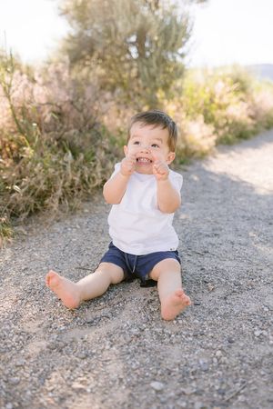 A small child sits on a dirt path making a playful expression while wearing a white shirt and navy shorts.