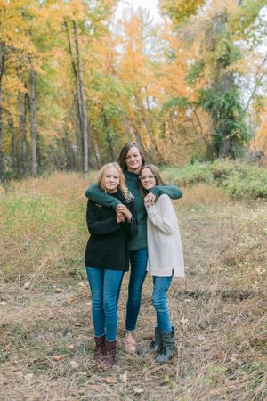 Three people embrace while standing in a wooded area with golden autumn foliage.