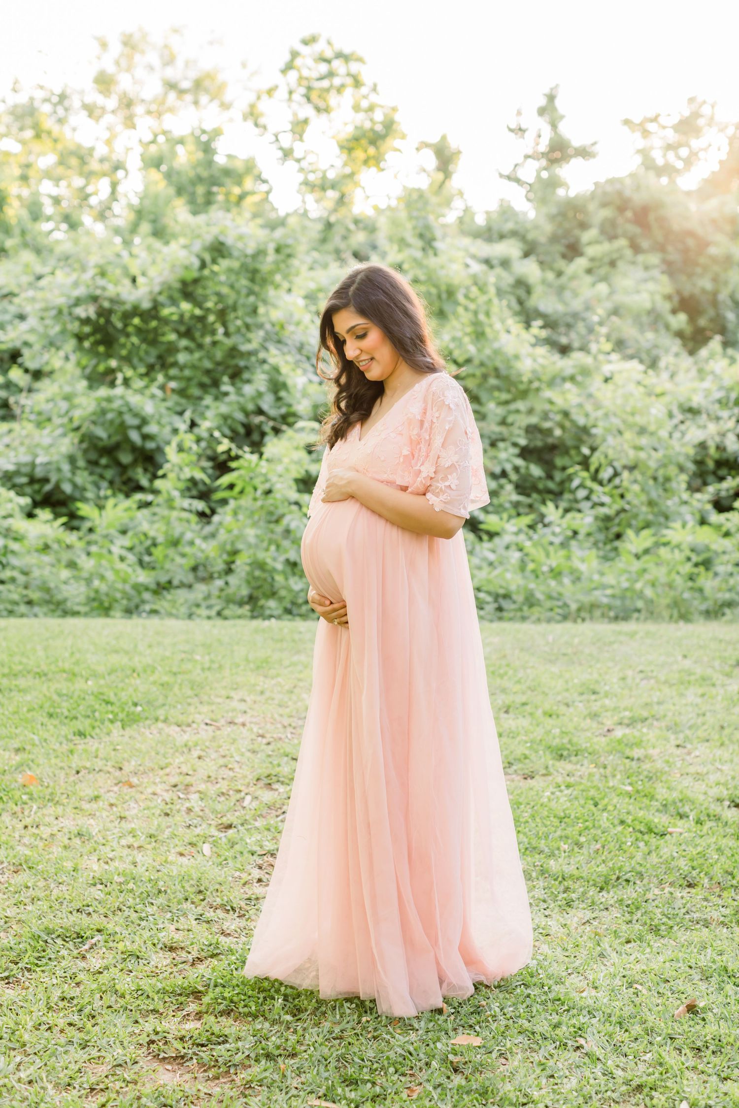 An expectant mother in a flowing pink dress stands in a garden setting during golden hour.