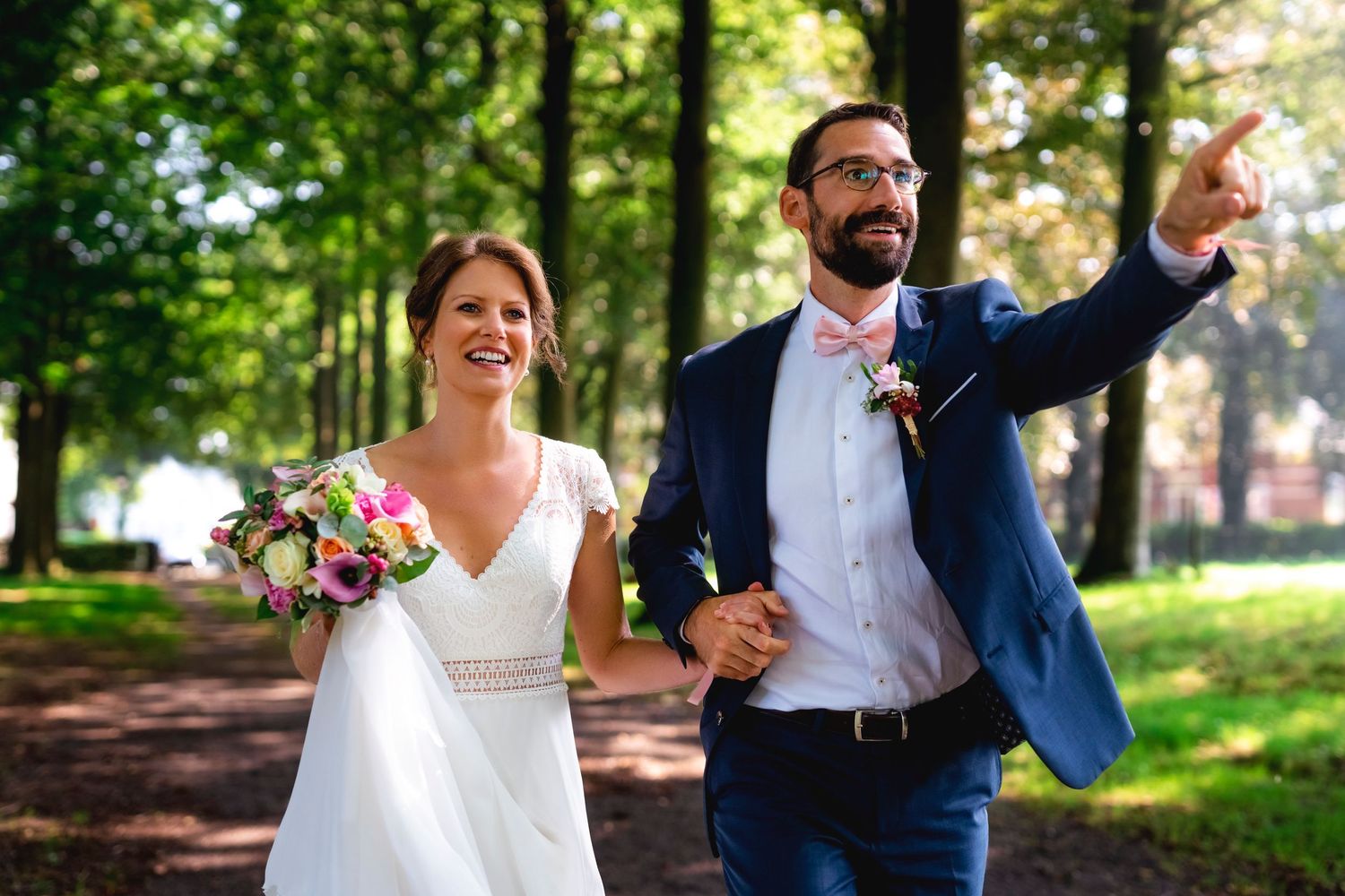 Couple strolls through park with bride holding pink bouquet as groom gestures ahead.