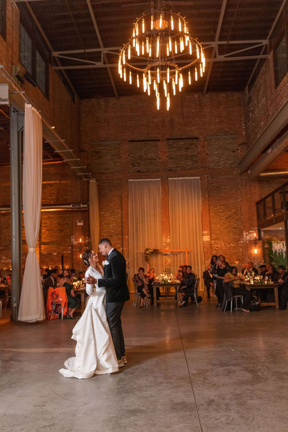 Elegant wedding dance under a dramatic chandelier in an industrial-style venue.
