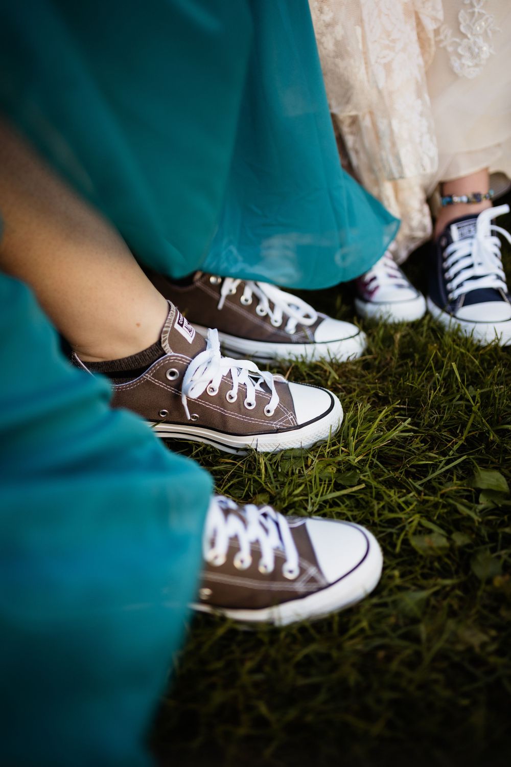 Brown Converse sneakers worn with teal dresses stand in grass at a wedding.