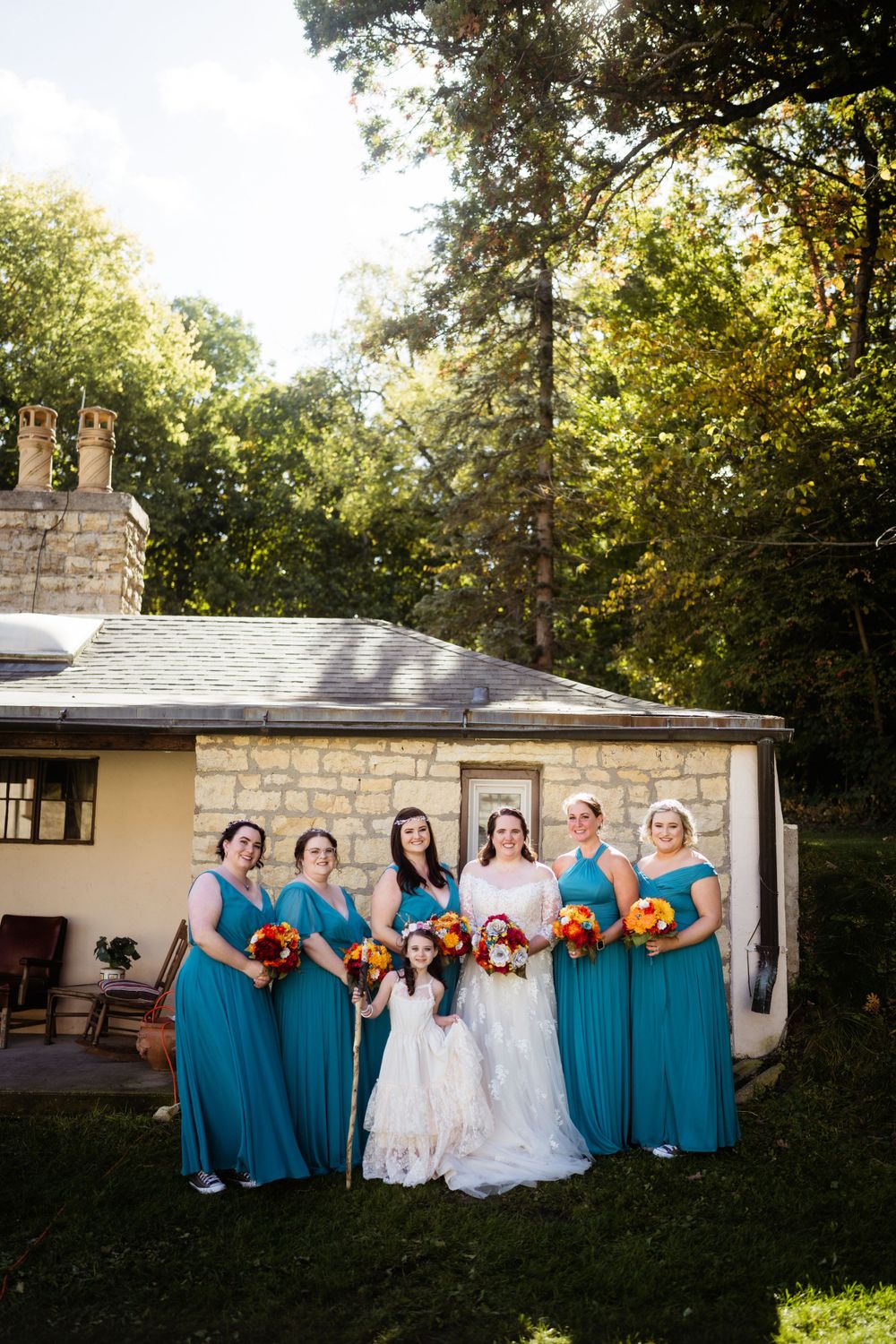 Wedding party poses in bright teal dresses holding orange and yellow bouquets against stone building.