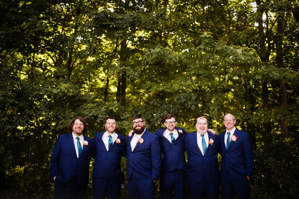 Groomsmen in matching navy blue suits pose together in front of trees.