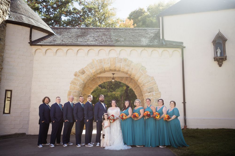 A wedding party poses in front of a stone arch wearing navy and teal colored attire.