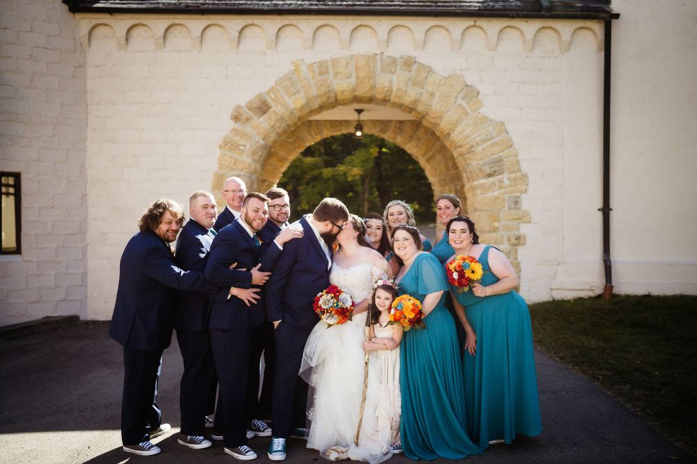Wedding party poses for a joyful group photo under a stone archway entrance.