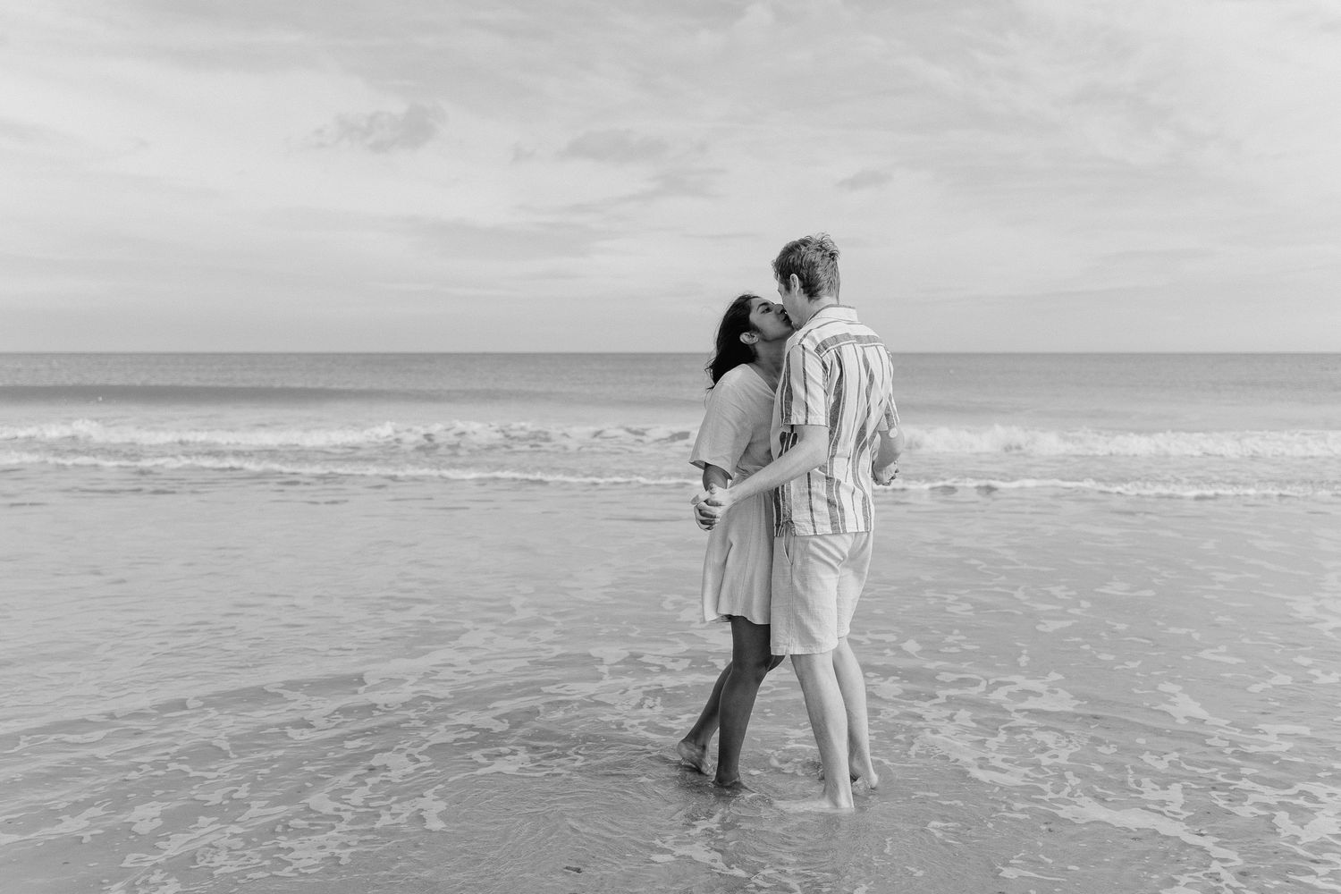 a couple are stood in the edge of the sea with water washing over their feet. they are chest to chest and kissing each other.