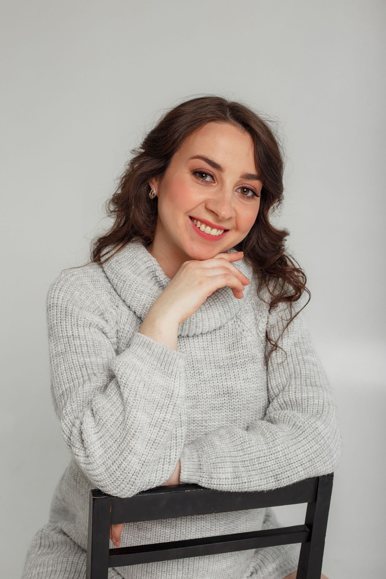 A smiling brunette woman in a gray turtleneck sweater poses gracefully on a black chair.
