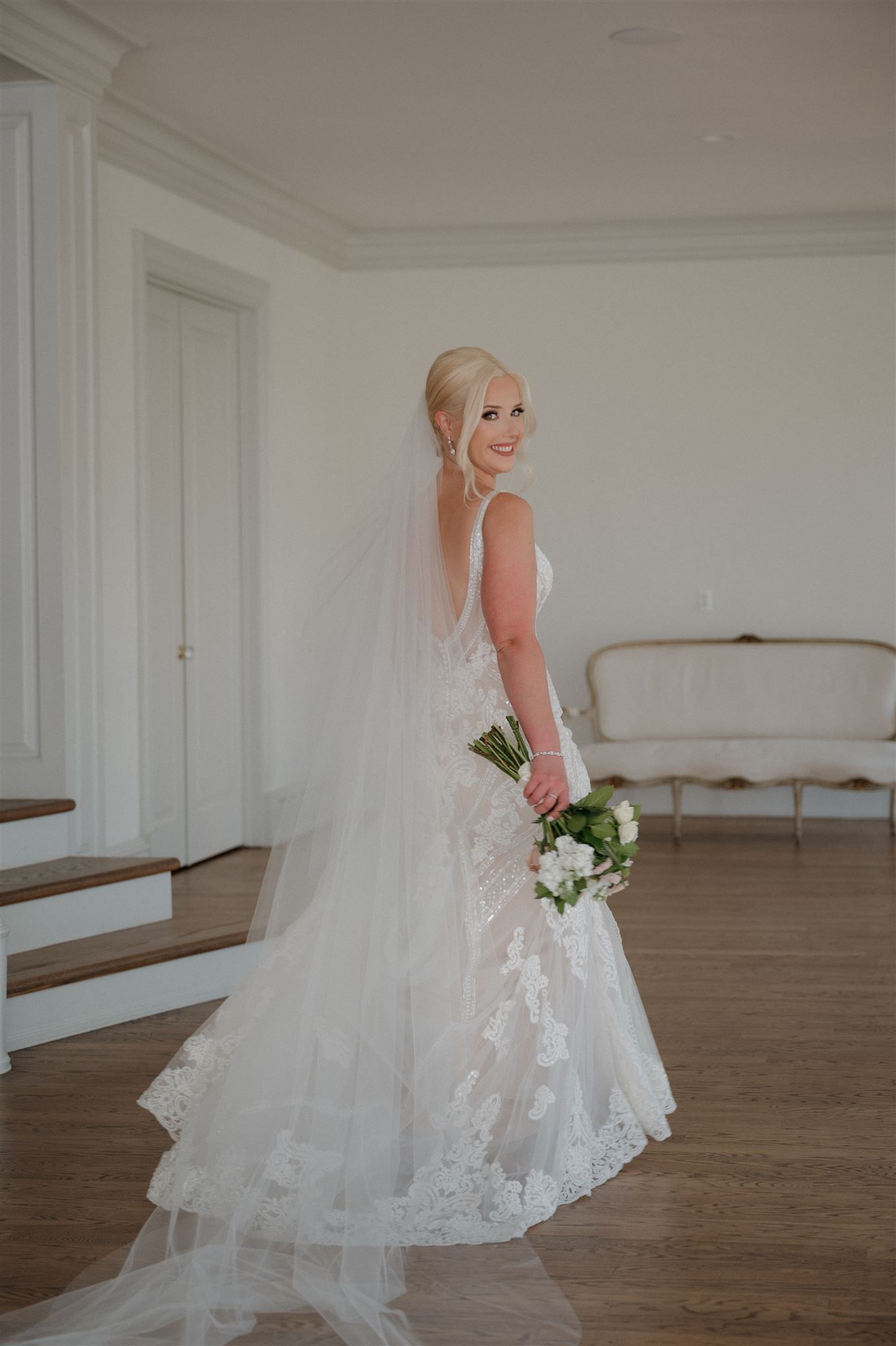 A bride in a lace wedding gown poses gracefully in an elegant room with hardwood floors.