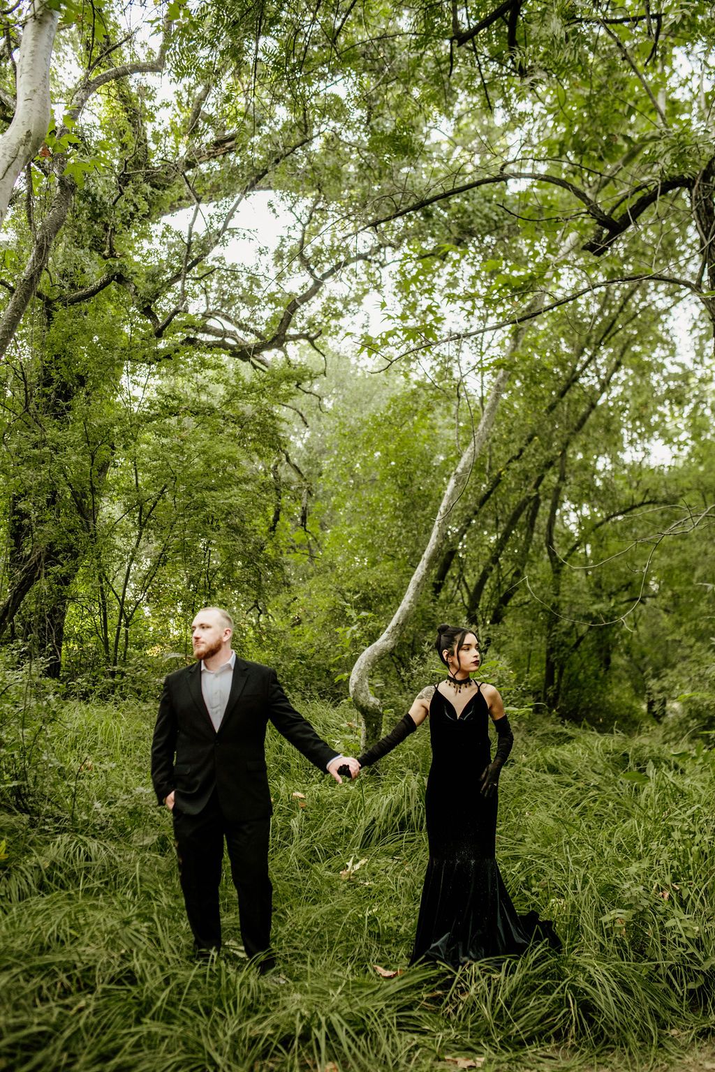 Couple in formal black attire hold hands while standing in lush green forest setting.