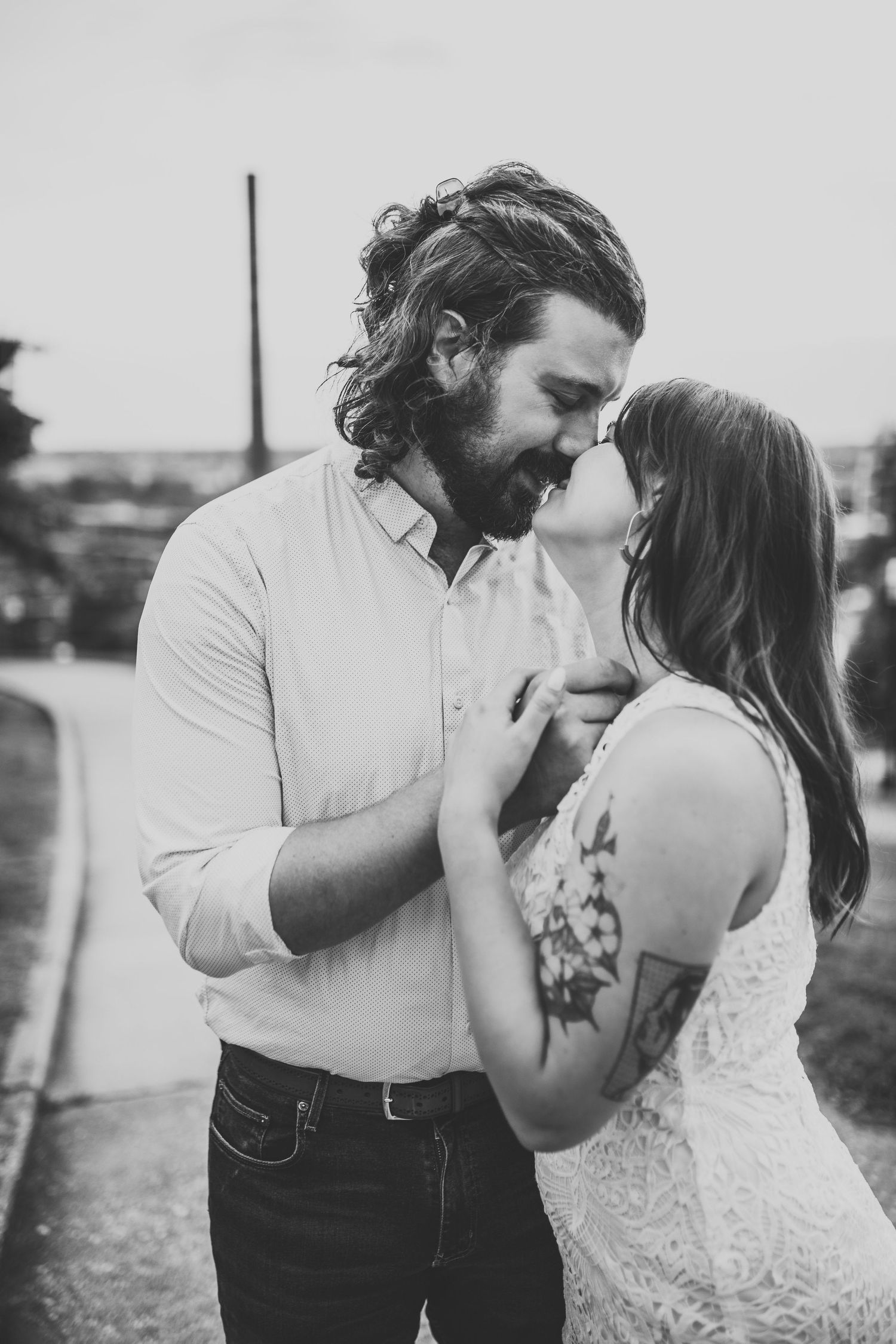 Black and white portrait of bearded man and tattooed woman sharing a romantic moment outdoors.