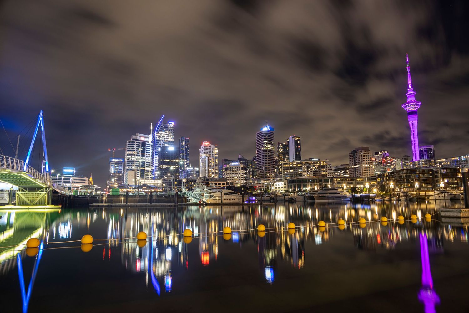 The Sky Tower and Viaduct Harbour shimmer in vibrant pinks and blues under a moody night sky.