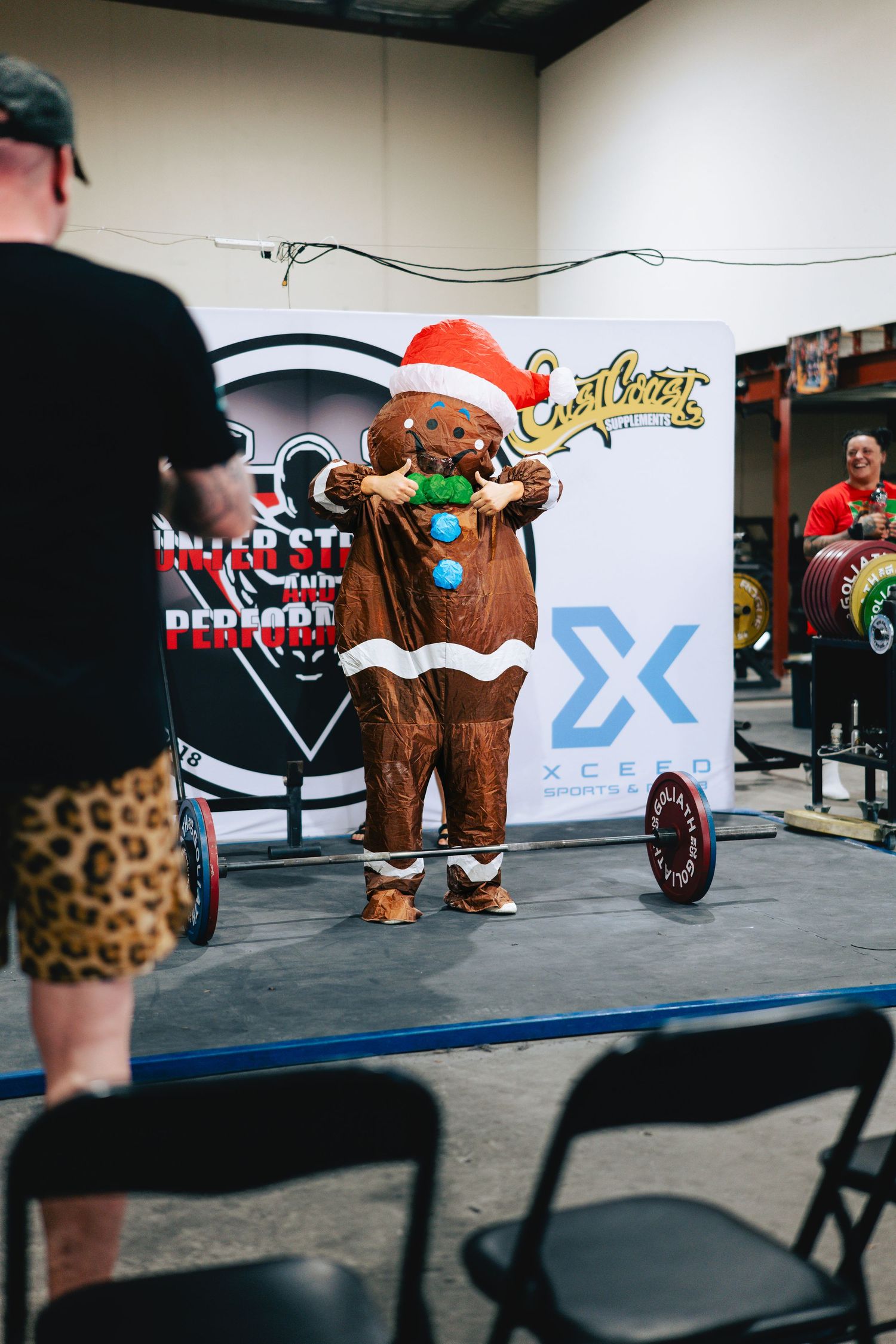 Person in gingerbread costume poses near fitness equipment during holiday themed gym event.