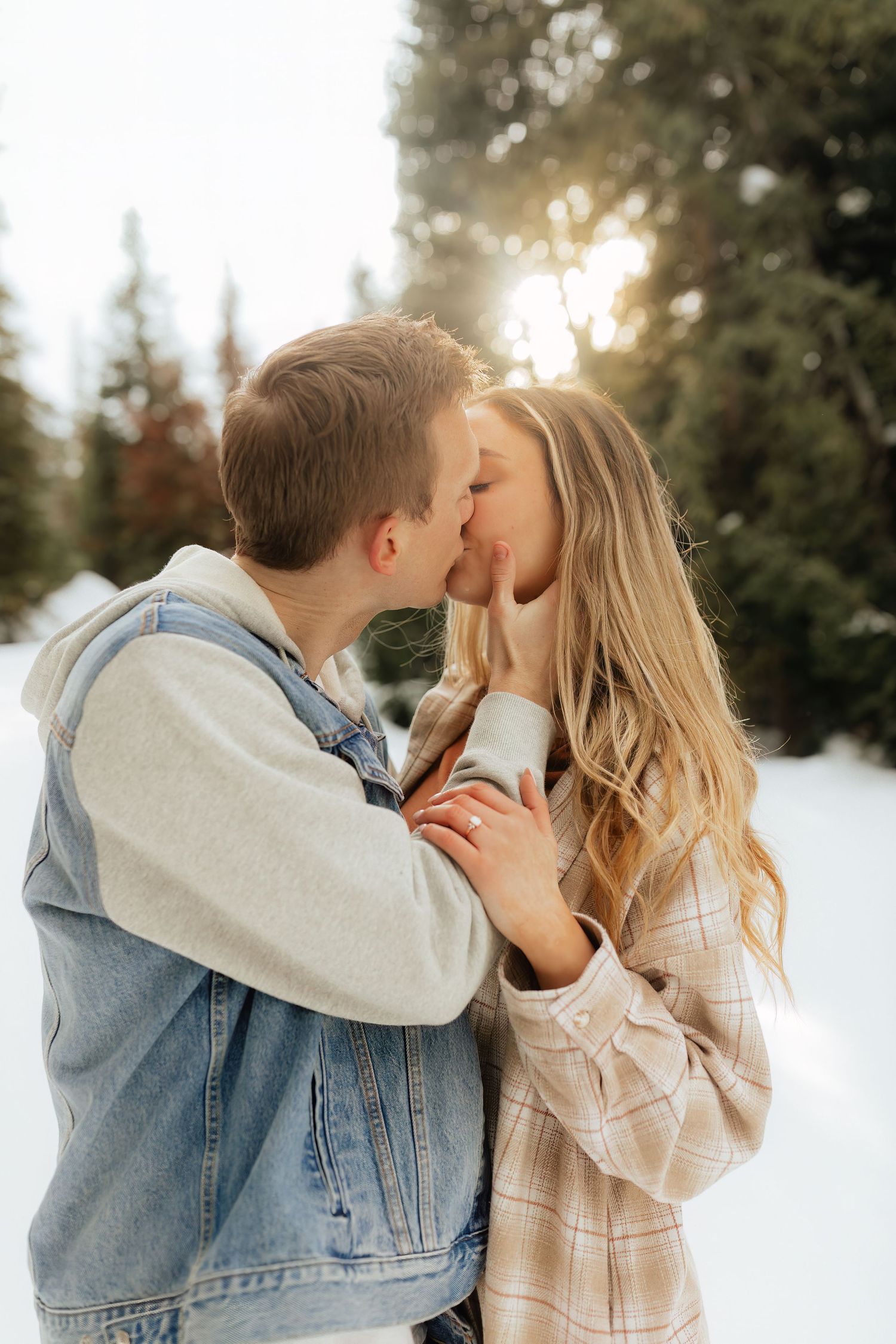 Woman and man kissing in winter pine trees during engagement photoshoot