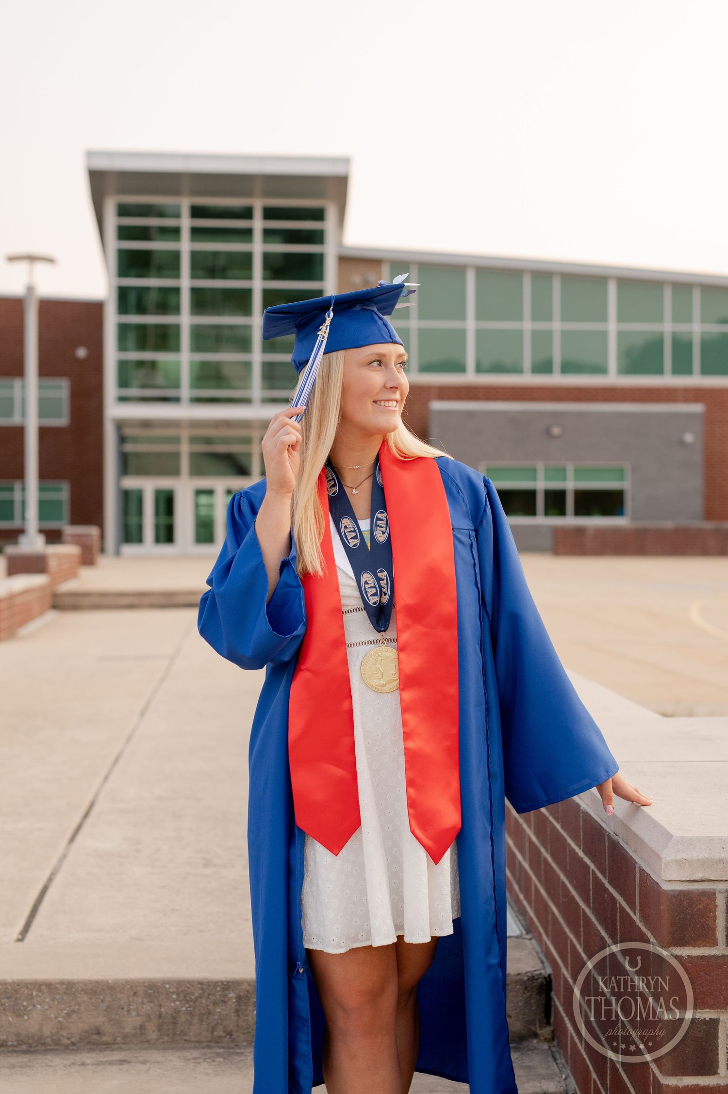 Graduate in blue cap and gown with red tie stands smiling outside modern campus building.