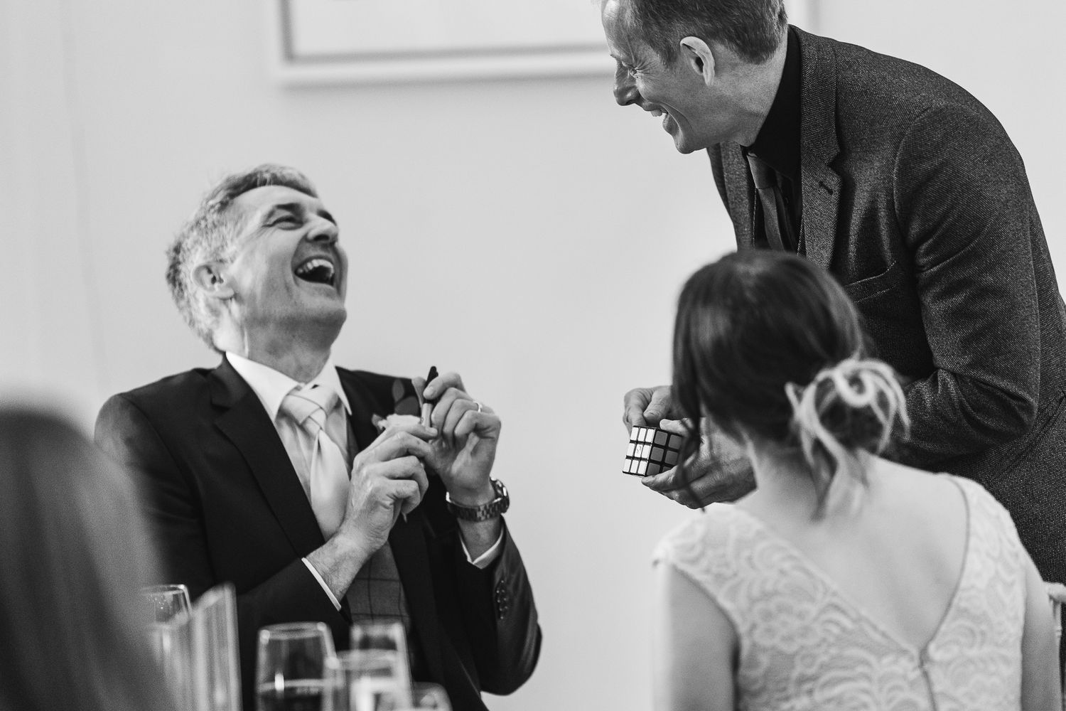Candid black and white photo captures genuine laughter during wedding reception toasts.