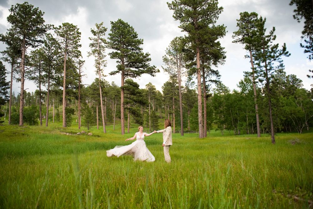 White dresses flowing in green meadow surrounded by tall pine trees.