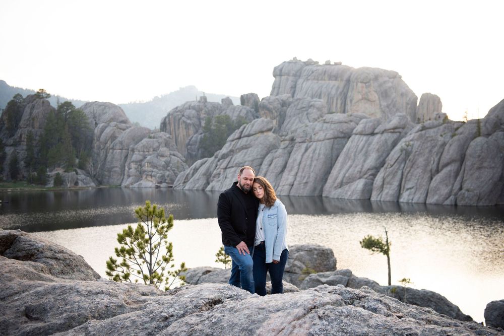 A couple poses on rocky cliffs overlooking a serene Sylvan lake at sunset.