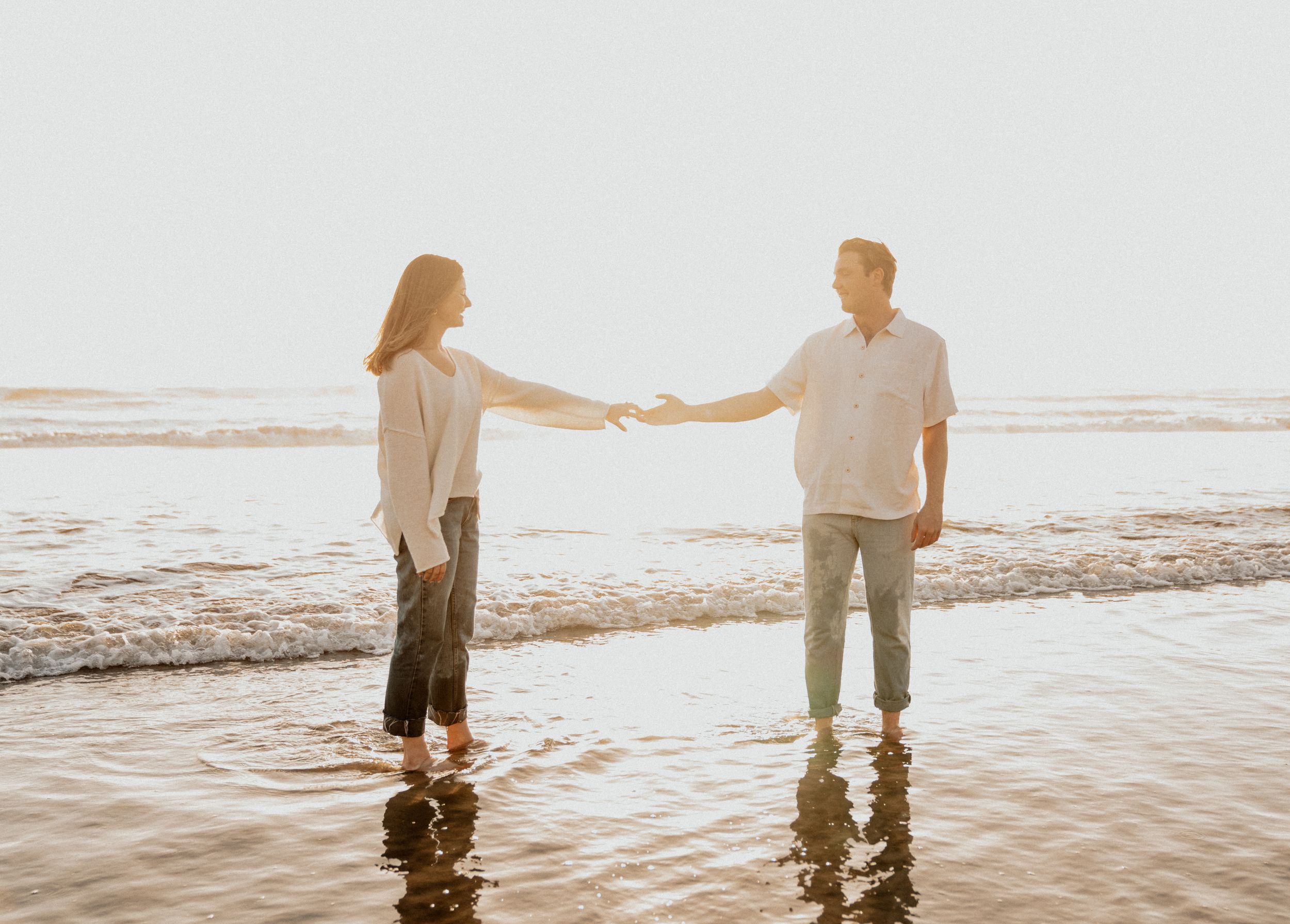 Two people hold hands while standing in shallow ocean water at sunset.