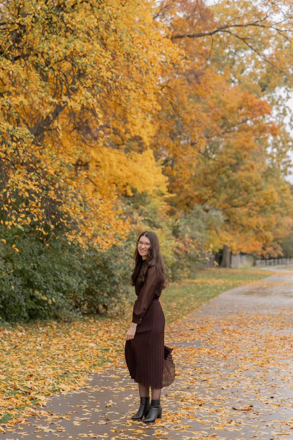 teenage girl in a brown dress standing on a walking path under a bright orange autumn tree in troy michigan