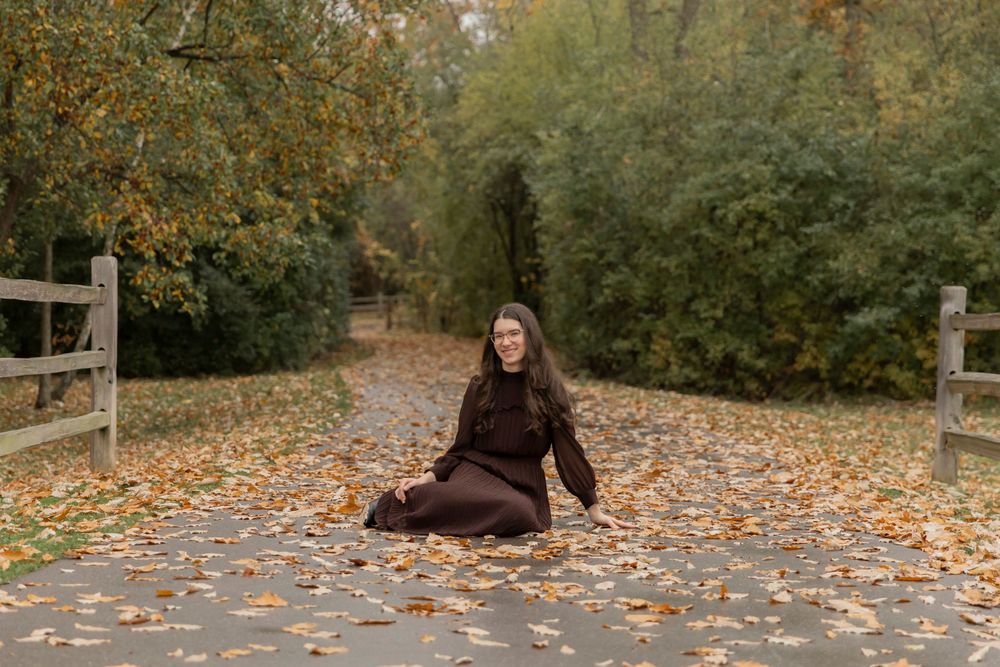 teenage girl wearing a brown dress sitting on a walking path at sylvan glen lake park