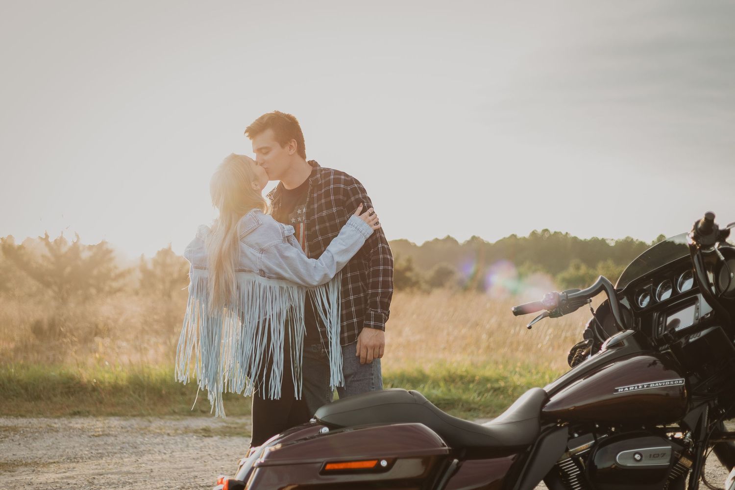 A romantic sunset moment beside a parked Harley Davidson motorcycle in a rural setting.