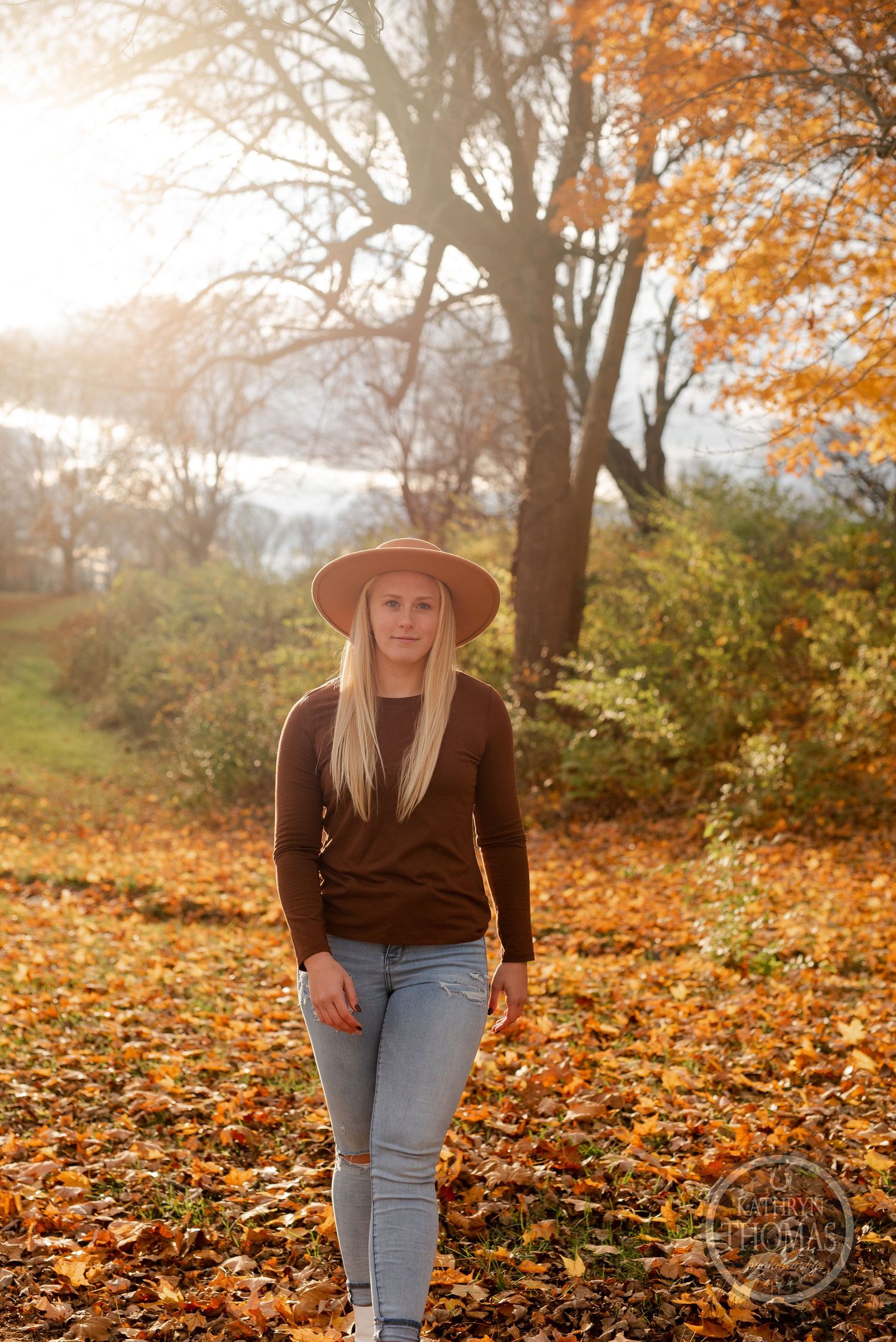 Person in brown sweater and hat walks through golden autumn landscape at sunset.