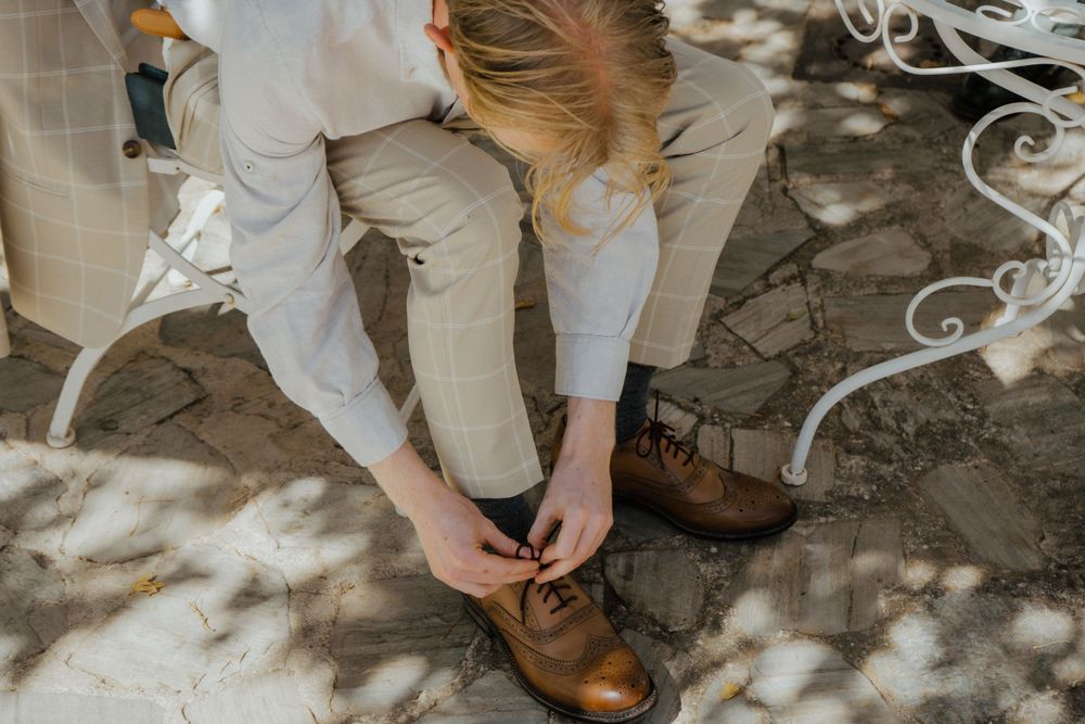 Person tying brown leather dress shoes while sitting on white iron garden chair. Destination Wedding in Malaga, Spain.