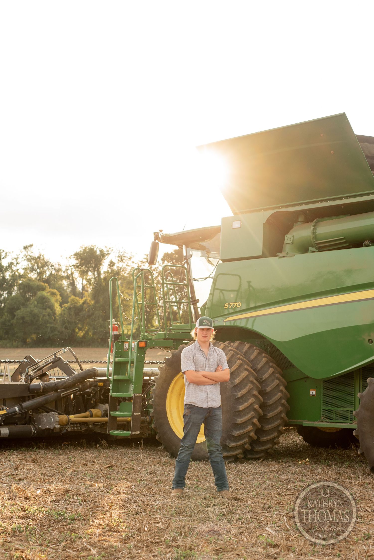 Farmer stands with crossed arms in front of combine harvester during sunset.