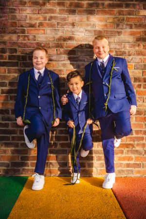 Three children stand together on colorful steps wearing matching royal blue suits and white sneakers against a brick wall.
