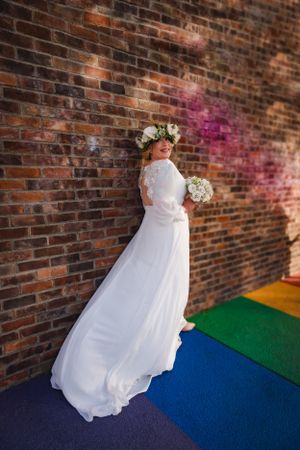 Bride in flowing white dress with floral crown stands against brick wall on vibrant rainbow walkway in Las Vegas' Arts District.