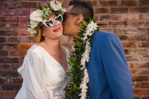 Elegant Vegas elopement couple wearing white dress and blue suit with floral leis embrace against brick wall backdrop.