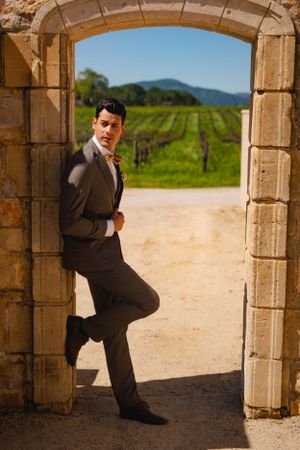 A groom poses in a stone archway overlooking scenic vineyard rows and mountains in the background.