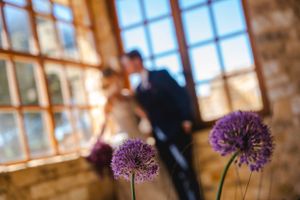 Purple allium flowers in foreground with soft focus wedding couple silhouette by window in rustic setting.