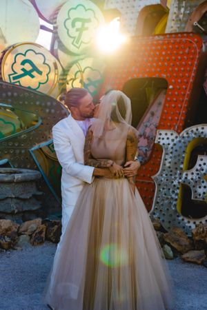 A couple in wedding attire embraces at the Neon Museum in front of a vintage Las Vegas sign with glowing lights at sunset.