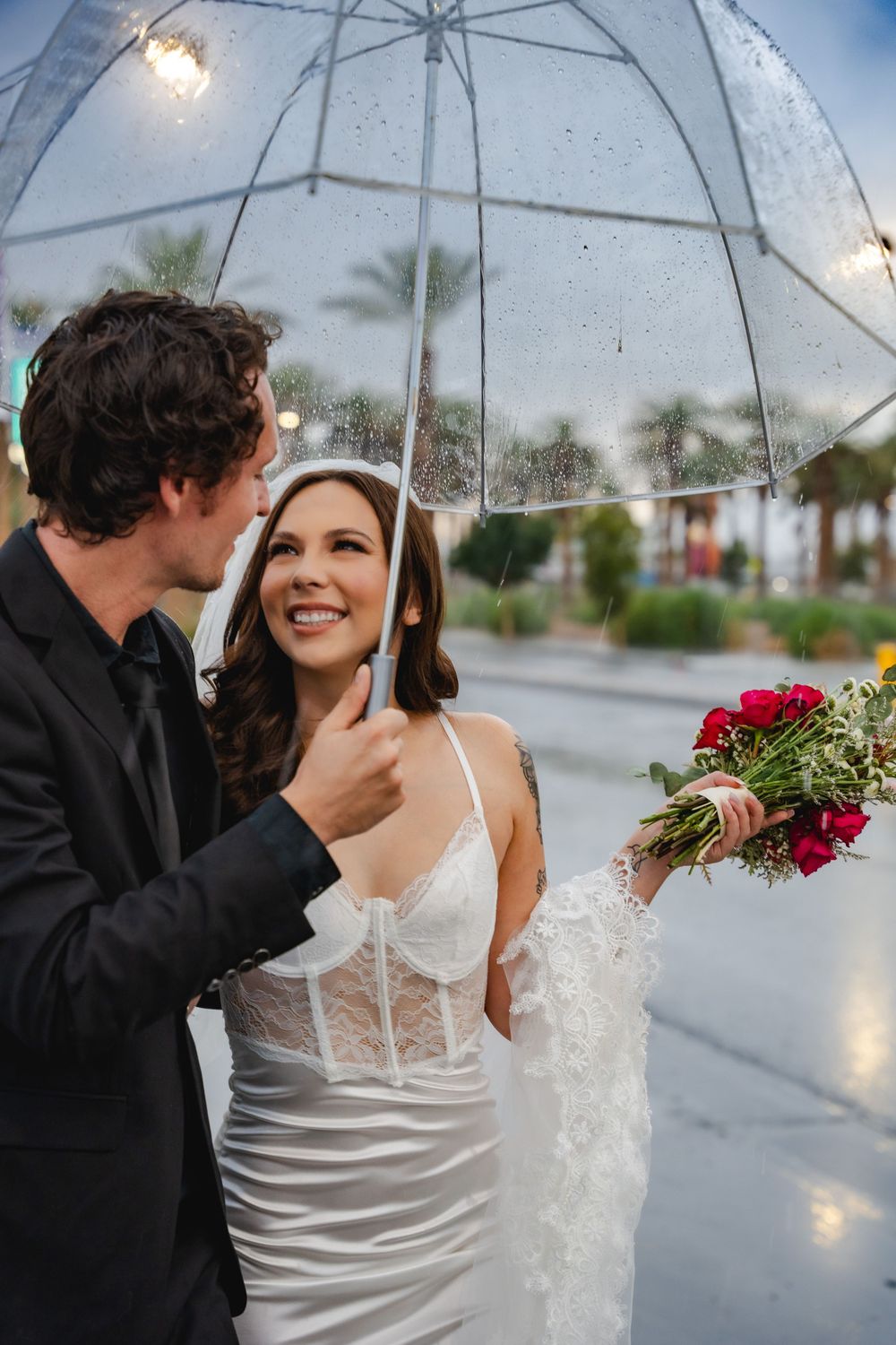 This Little White Chapel elopement couple shares a moment under a clear umbrella in the rain, with red roses and palm trees visible.