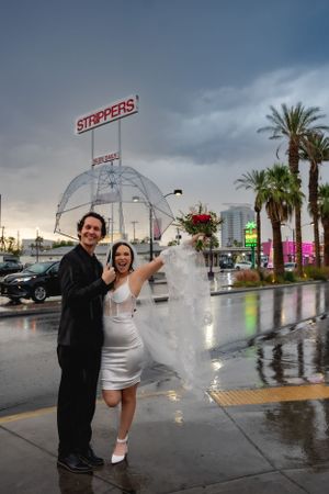 An elopement couple poses with an umbrella on a rainy Las Vegas evening with palm trees and neon signs in view.
