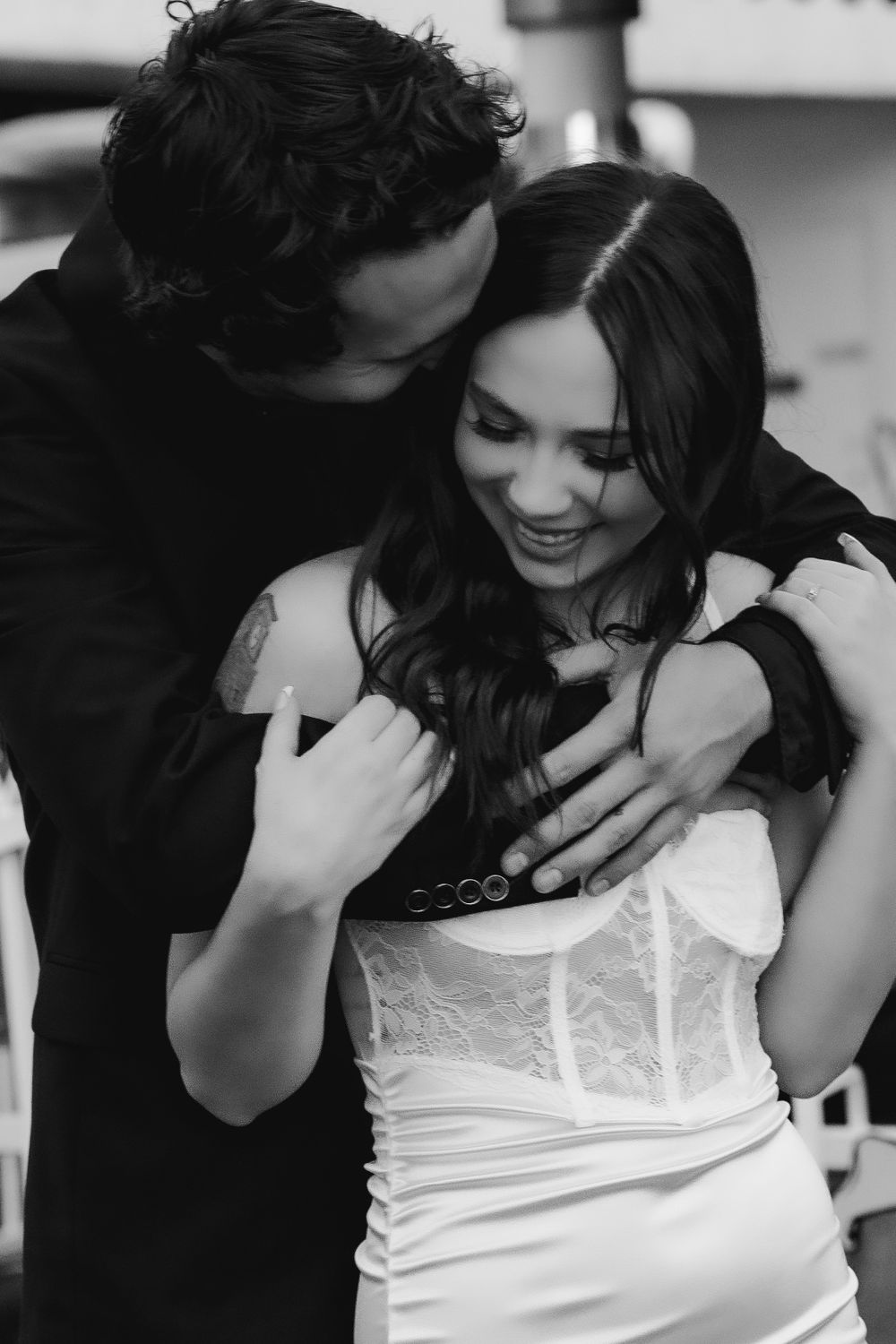 A romantic black and white photo of a destination wedding couple embracing, with the woman wearing a white lace dress and smiling.