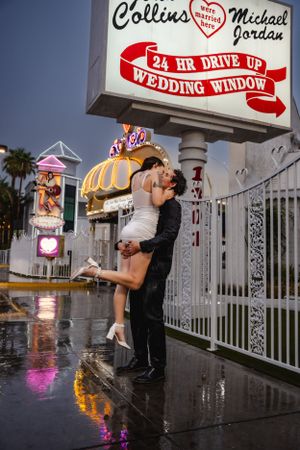 Couple shares romantic kiss under illuminated 24 hour drive-up wedding window sign in Las Vegas on rainy evening.