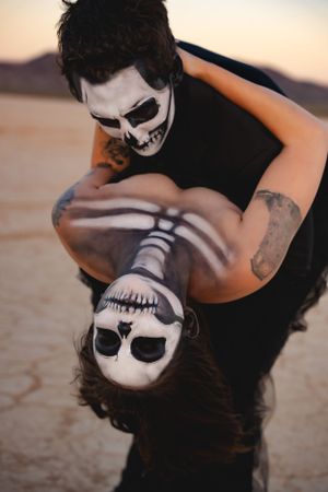 Two people with skeleton face paint embrace in artistic Halloween-themed portrait at sunset in desert landscape.