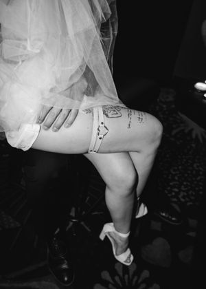 Black and white photo of leg with garter belt tattoo visible under white lace fabric and high heels.