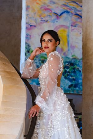 Elegant bridal gown with sheer sleeves and pearl embellishments shown against colorful abstract artwork background at the Sunstone Winery.
