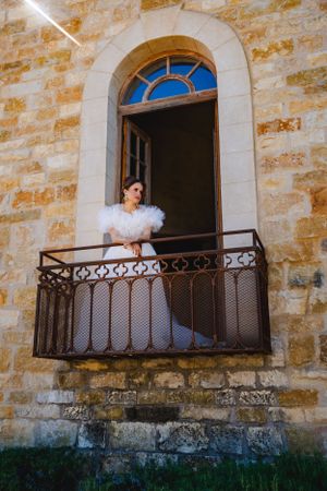 A bride in an ornate white dress stands on a vintage iron-railed balcony against weathered stone walls of the Sunstone Winery.