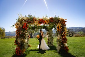Beautiful wedding couple under floral wedding arch with orange and red flowers overlooking mountain landscape at Sunstone Winery.