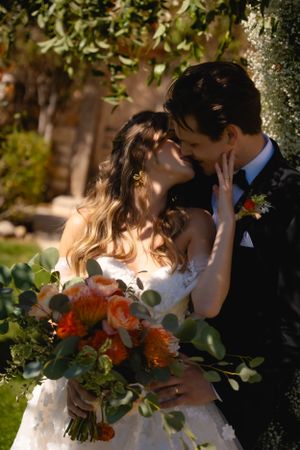 A romantic kiss between newlyweds in a garden setting with a vibrant orange and green floral bouquet.