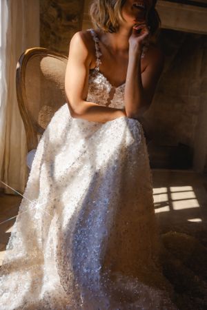 A beautiful white lace wedding dress sparkles in sunlight streaming through a window as the wearer sits in an antique chair.