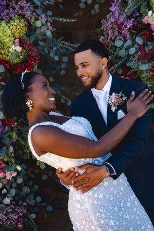 A joyful wedding couple embraces in front of a vibrant floral backdrop with purple, red and green blooms.